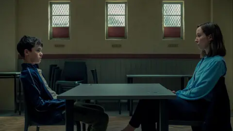 Ben Blackall/Netflix Still of Owen Cooper as Jamie Miller and Erin Doherty as Briony Ariston in the Adolescence drama. They sit opposite each other with a table in between in a school room with chairs and tables stacked against a wall with three windows.