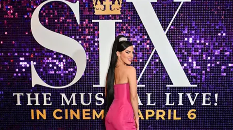 A woman with a long dark ponytail and pink dress is smiling in front of the logo for Six the Musical
