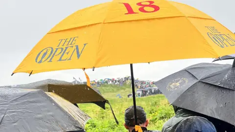 Os fãs de golfe estão fora de seus guarda -chuvas e para ver as aberturas abertas durante a chuva. O curso mostra vários guarda -chuvas encharcados e situações úmidas no curso.