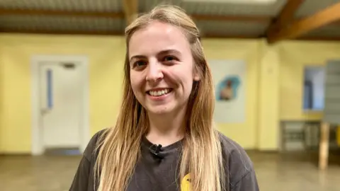 Abi Tate, who has long blonde hair and is wearing a dark grey T-shirt, smiling at the camera inside a large room at the Dogs Trust in Salisbury. The walls are painted bright yellow, the floor is concrete and the ceiling is corrugated metal panelling.