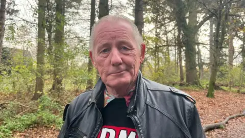 Ellen Knight/BBC A man with swept-back white hair looking into the camera with a solemn expression. He is wearing a black leather jacket with a black and red jumper on underneath. He is standing in woodland, with lots of trees with green, brown and orange leaves on the trees. The ground is covered in a layer of fallen leaves, and the sky is grey and overcast. 