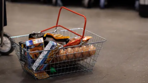 A shopping basket full of beige groceries sits on a grey concrete supermarket floor.