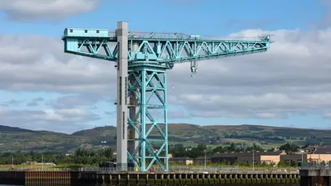 A large latticed steel crane with a lift shaft at itss side with hills in the background