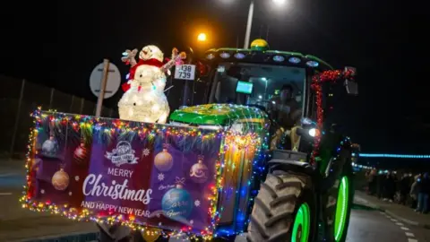 The front of a green tractor covered in Christmas lights. On the front there is a purple sign with the words 'Merry Christmas and Happy New Year' and a light-up snowman wearing a red scarf. In the distance to the right are crowds of people watching as the tractors drive down the road.