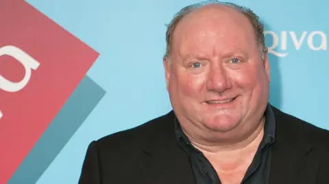 Alan Brazil wearing a black suit jacket and shirt at a press event. The backing behind him is turquoise blue.