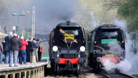 Two steam engines on tracks with crowds of people watching and taking photographs
