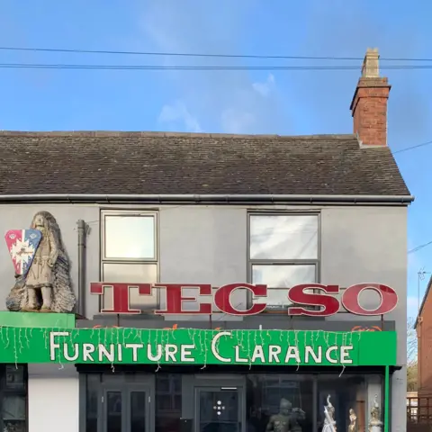 Tom Hicks A shop stands beneath a clear blue sky. The shop name is Tecso - in the same red type used by supermarket Tesco. Underneath the name, in white lettering on a green background, is spelled: Furniture Clarance.