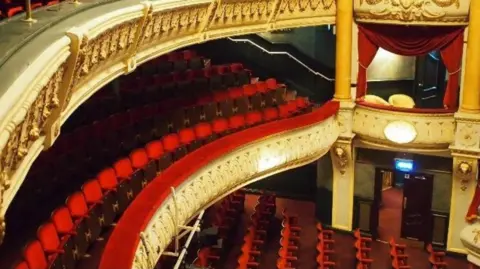 A historic theatre with red velvet seating, gold detailing, and tiered balconies facing the stage.