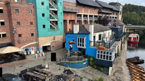 LDRS Another view of the pub and the mural which is in front of a large brick building with a turquoise wall. There is a river in front of the pub with rowing boats tied to the side. There is also a beer garden to the left of the building.