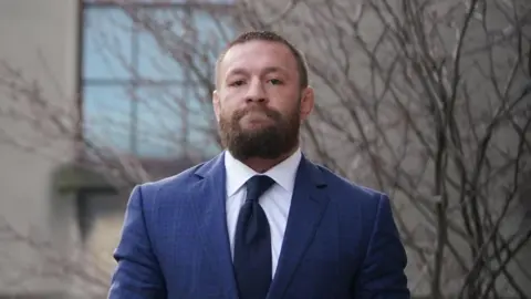 PA Media Conor McGregor, with short brown hair and a brown beard, wearing a navy suit, white shirt and navy tie
