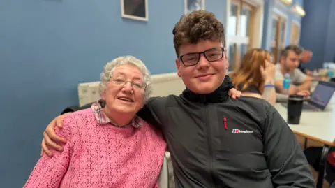 BBC/Jim Scott Veterans Trust volunteer Luke Underwood, right, has his arm around Ruth Bullen, who is wearing a pink jumper on top of a tartan patterned shirt. They have just finished playing a round of Dominoes. 