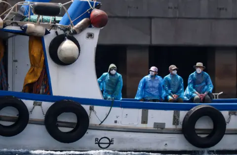Four people are sitting on the back of a white boat with blue trim, and tyres hanging alongside. The four people are wearing blue protective raincoats and white facemasks.