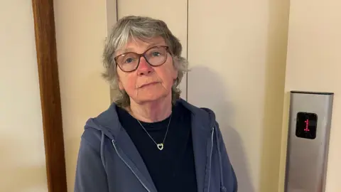 The image shows a person standing in front of a lift door. The lady is wearing a glasses, a dark blue hoodie and a black shirt underneath, along with a necklace that has a heart-shaped pendant. There is an elevator control panel with the number "1" illuminated in red.