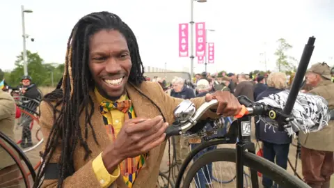 A man in a beige blazer, yellow shirt, patterned waistcoat and matching bow tie holds the handlebars of a Penny Farthing. People stand in the background.