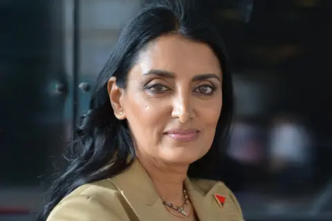 Aneeta Prem Aneeta Prem looks directly at the camera. She has dark hair and is wearing a pale coloured suit jacket. There is a red and black badge on her lapel in the shape of a triangle. 