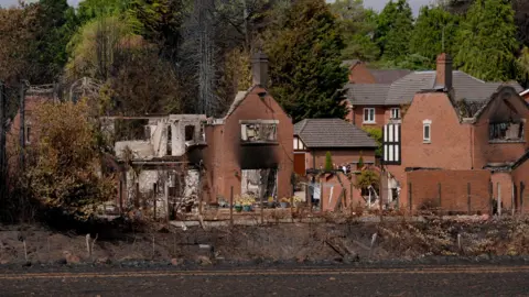 PA Media A distant photo of several houses completely destroyed by fire. They do not have roofs and some walls have fallen away. they are blackened with smoke 