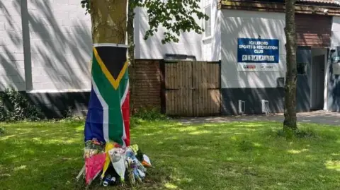 Flowers left at the bottom of a tree outside a white building, which has a blue sign that reads:  "Ickleford Sports & Recreation Club". South Africa's national flag is hung from the tree above the flowers. 