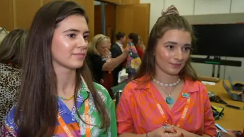 A woman in a brightly coloured shirt with dark hair and another woman with lighter hair and a pink and orange shirt stand in a busy room in Westminster. 