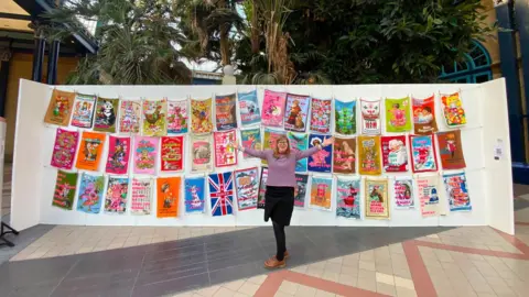 Large display of colourful tea towel artworks hung on lines, with one person standing in front of the installation.