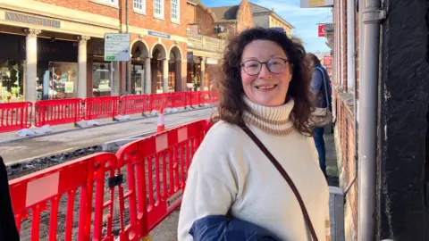 Miranda Shawcross, a middle-aged woman with long brown hair and glasses, wearing a white polar neck sweater, stands in Farnham in front of a set of red plastic railings guarding roadworks.