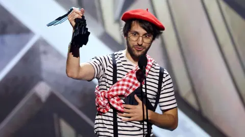 AFP via Getty Images Guillaume Broche, wearing a red beret, striped t-shirt plus red and white checked sash around his shoulder. He's standing behind two microphones, delivering a speech to the audience.