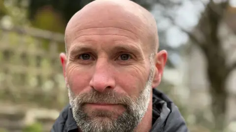 A close up photo of a man stood outside. The man is bald with a brown and grey beard and is wearing a black coat. There is a tree out of focus in the background, along with other greenery. 