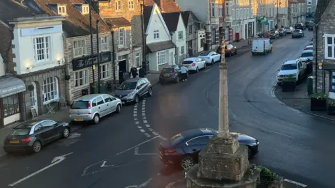 A colour photograph matching the exact location of the black and white version above, but taken in January 2025 - the buildings and war memorial remain as modern cars pass by.