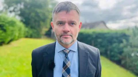 Det Insp James Callander looks into the camera, with green grass and bushes behind him