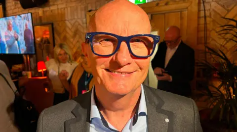 A man in a grey suit with a blue shirt. He is wearing blue glasses. There is a room with people behind him.
