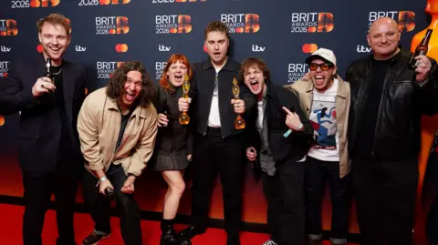 Ian West/PA Wire Four men and one woman dressed in smart dark clothing and two men wearing beige jackets celebrating Sam Fender's win on the red carpet. He is stood in the middle holding two awards. There is a Brit Awards 2026 backdrop behind them.