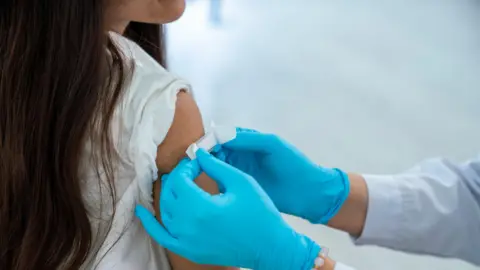 Getty Images A plaster is applied by a medical worker with blue gloves to the upper arm of a young girl after vaccination