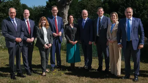 State of Guernsey Six white middle-aged men in dark blue or navy suits and three smartly dressed women standing together in a garden. They are all looking at the camera smiling. There are trees in the background and blue skies. 