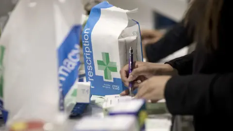 A stock image of a woman picking up a prescription at a Boots pharmacy. She is signing something with a purple pen and beside her on the counter is a blue and white bag containing her prescription. In the foreground, out of focus, there are various medications.