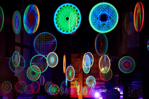 Matthew Andrews Dozens of wheels are suspended from the air and illuminated in different neon colours.