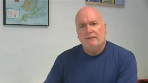 Language school principal Lee Shutler faces the camera looking serious as he is interviewed by the BBC, with a map on the wall behind him