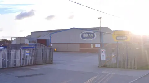 A wide picture of the Nolan Recycling warehouse unit behind open grey gates. The sun is shining in the background of the image.