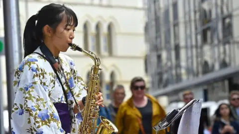 A girl wearing a white kimono with a pattern of blue flowers and green leaves plays a saxophone. In the background, people are walking along a city street. 
