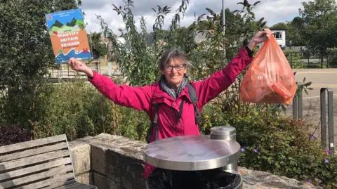 Ruth Major wearing a bright pink raincoat and grey rucksack. She is holding up an orange Sainsbury's plastic bag full of collected litter in one hand, and a campaign card in the other.