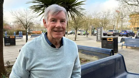 Martin Heath/BBC Sam Crooks with short silver hair smiling at the camera and wearing a dark blue shirt and light blue sweater. He is standing outside Centre MK in front of metal seats on paving slabs, with street furniture including litter bins and trees.