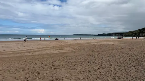 A wide shot of Whiterocks beach, people can be seen in the distance under a cloudy sky with some blue.