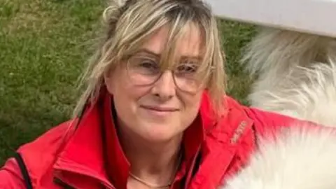 A close up of a woman with blonde hair tied behind her with strands across her forehead wearing a red coat with jacket collars sticking up