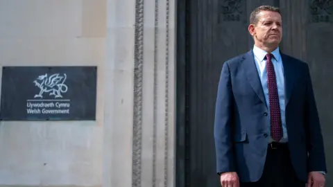 Wales First Minister, Rhun ap Iorwerth wearing a navy suit, light blue shirt and maroon tie with white spots. He's standing outside an old building with white stone walls and large wooden door. On the wall is slate plaque with "Welsh Government Llywodraeth Cymru" written on it. 