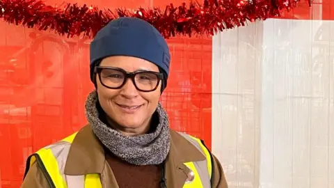 Golda Rosheuvel wearing an brown coat and faux fur grey scarf with a high vis tabard and a blue beanie-type hat, standing in front or some red tinsel.