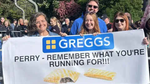 BBC/Eleanor Ingleby Four people hold up a large sign with the Greggs logo on it. Beneath it, the sign reads: Perry - remember what you're running for, and below that, there is a picture of a pasty and a sausage roll.