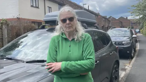 A woman with white hair wearing a green jumper. Her arms are folded. She is wearing glasses and is looking into the camera. There are two parked cars behind her.