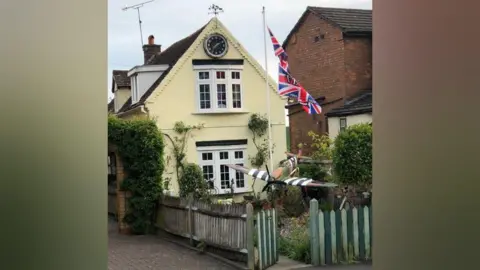 Ray Burden A wide shot of the plane in front of a cream house. There is a white flag pole with union jack flags that read "VE Day". The fence around the garden is green, and there is a black clock on the house.