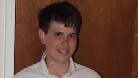 Jimmy Mizen, a boy with black hair wearing a white shirt, smiles at the camera against a brown background.