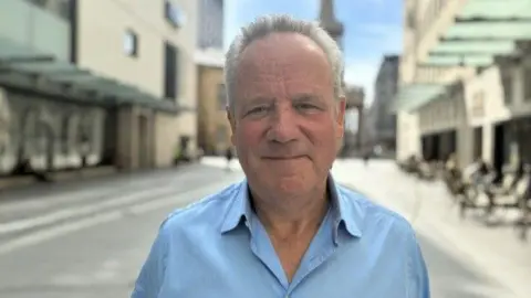Chris wears a light blue collared shirt and has a mild smile on his face, while standing outside on a sunny day surounded by buildings.