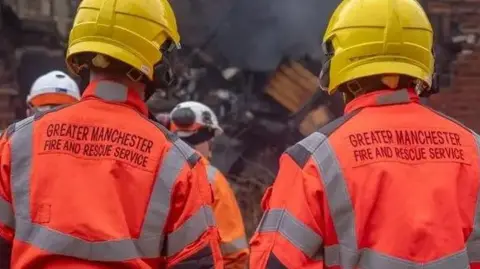 Two Greater Manchester Fire and Rescue Service firefighters in orange safety jackets and yellow hard-hats looking at the scene of a fire. 