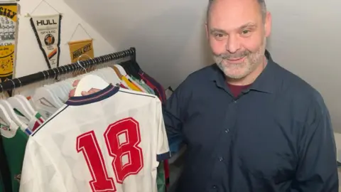 An man with cropped grey hair and stubble holds an original white England shirt from 1988, with the number 18 on the back, on a hanger. There is a rail of shirts behind him and Hull City pennants hanging on the wall. He is wearing a navy coloured short with a burgundy undershirt 